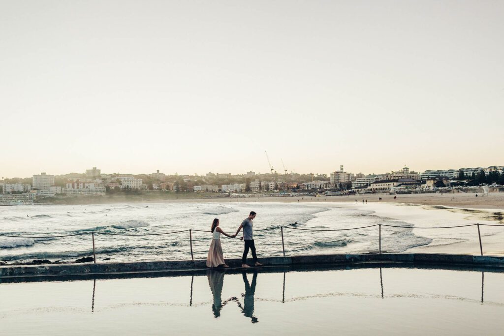 A male and a female are holding hands walking along the edge of an ocean pool at Bodhi beach the the sun setting the the background,