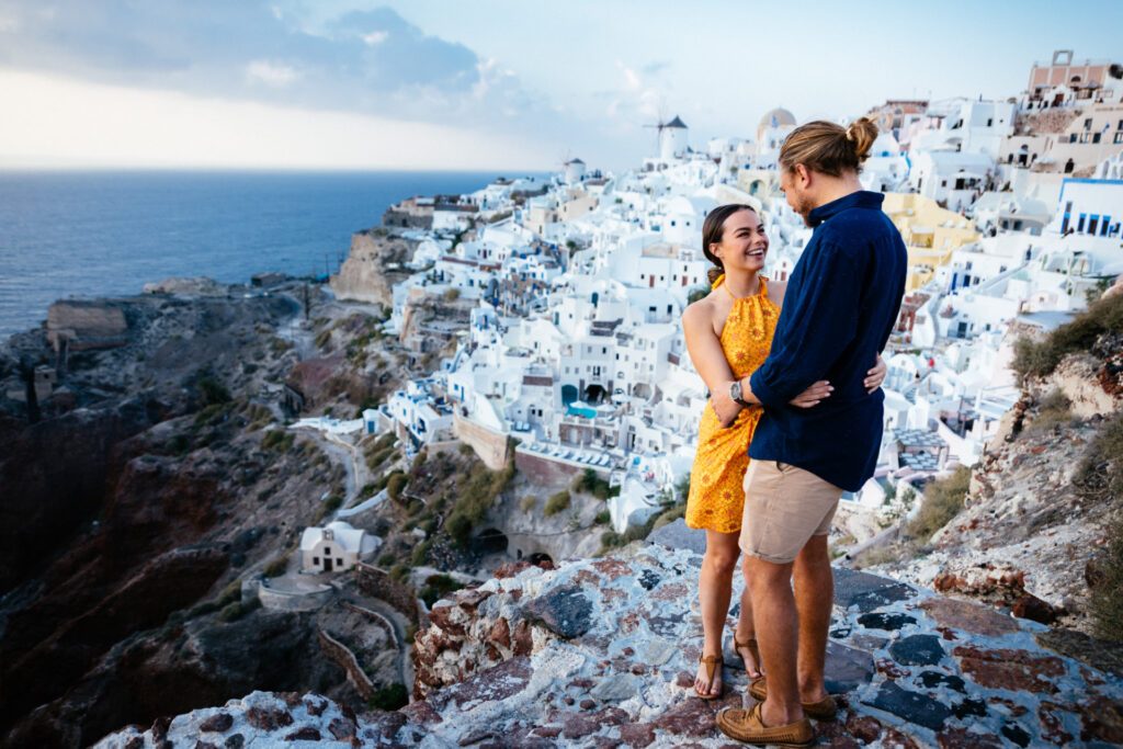 A female in a yellow dress is hugging a man in a blue shirt with the white houses ofSantorini and the ocean in the background