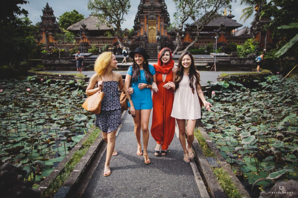 Four females are walking in front of a temple in Bali all linking arms and smiling and have their photo captured by a travel photographer.