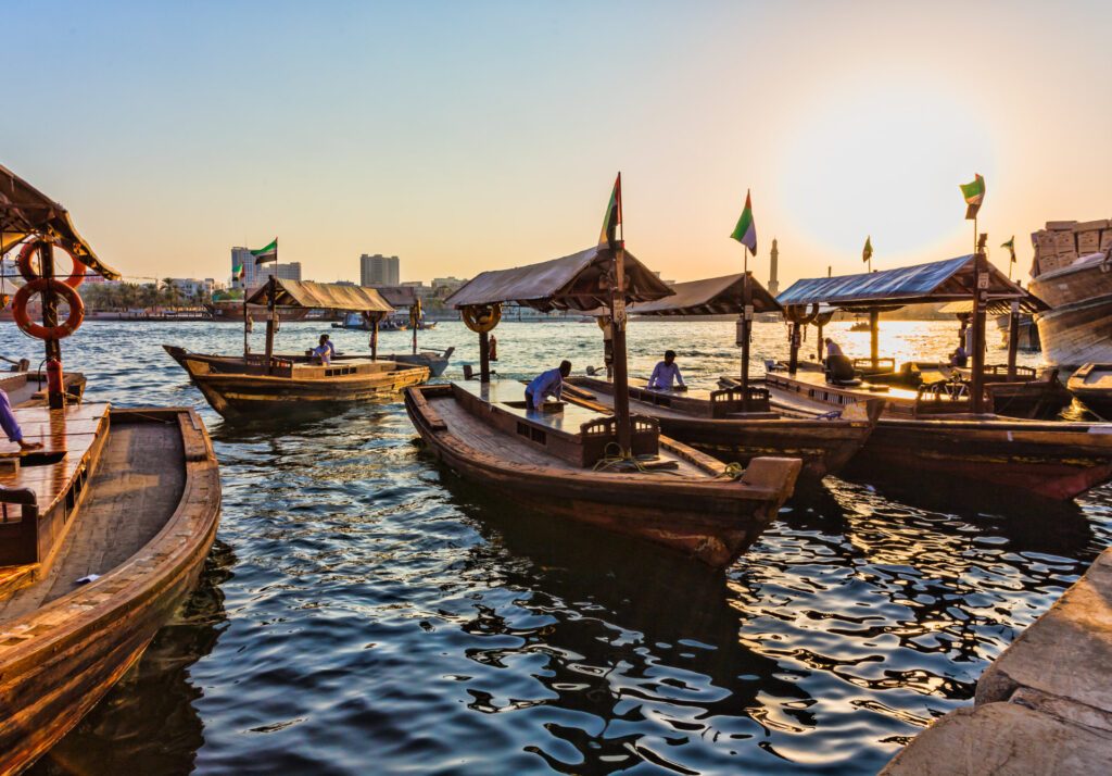 Traditional abra boats in Old Dubai floating along the river with the sun setting in the background - Luxury Escapes