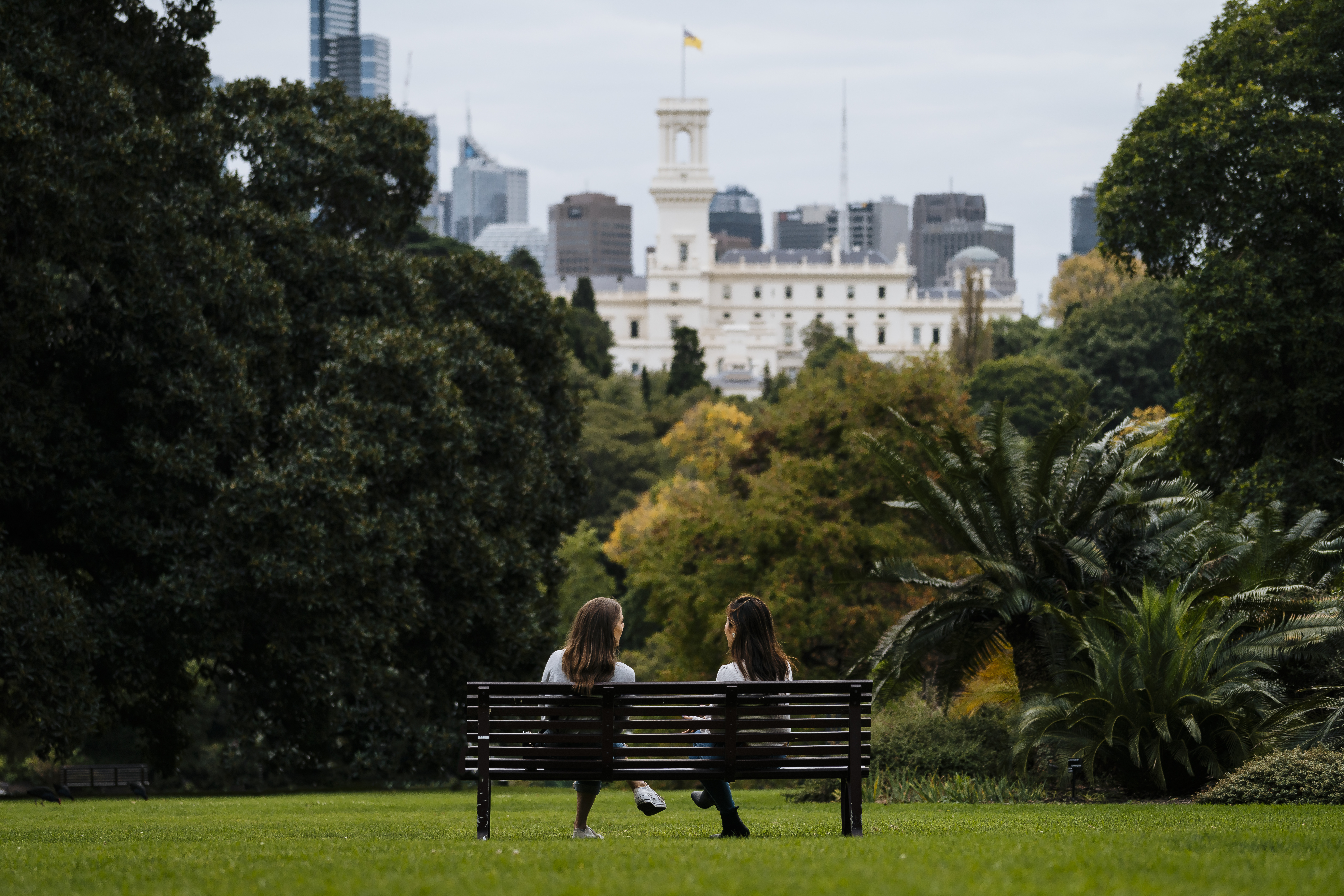 Two people sitting on a bench in Melbourne Royal Botanical Gardens with the CBD visible in the background.|||||