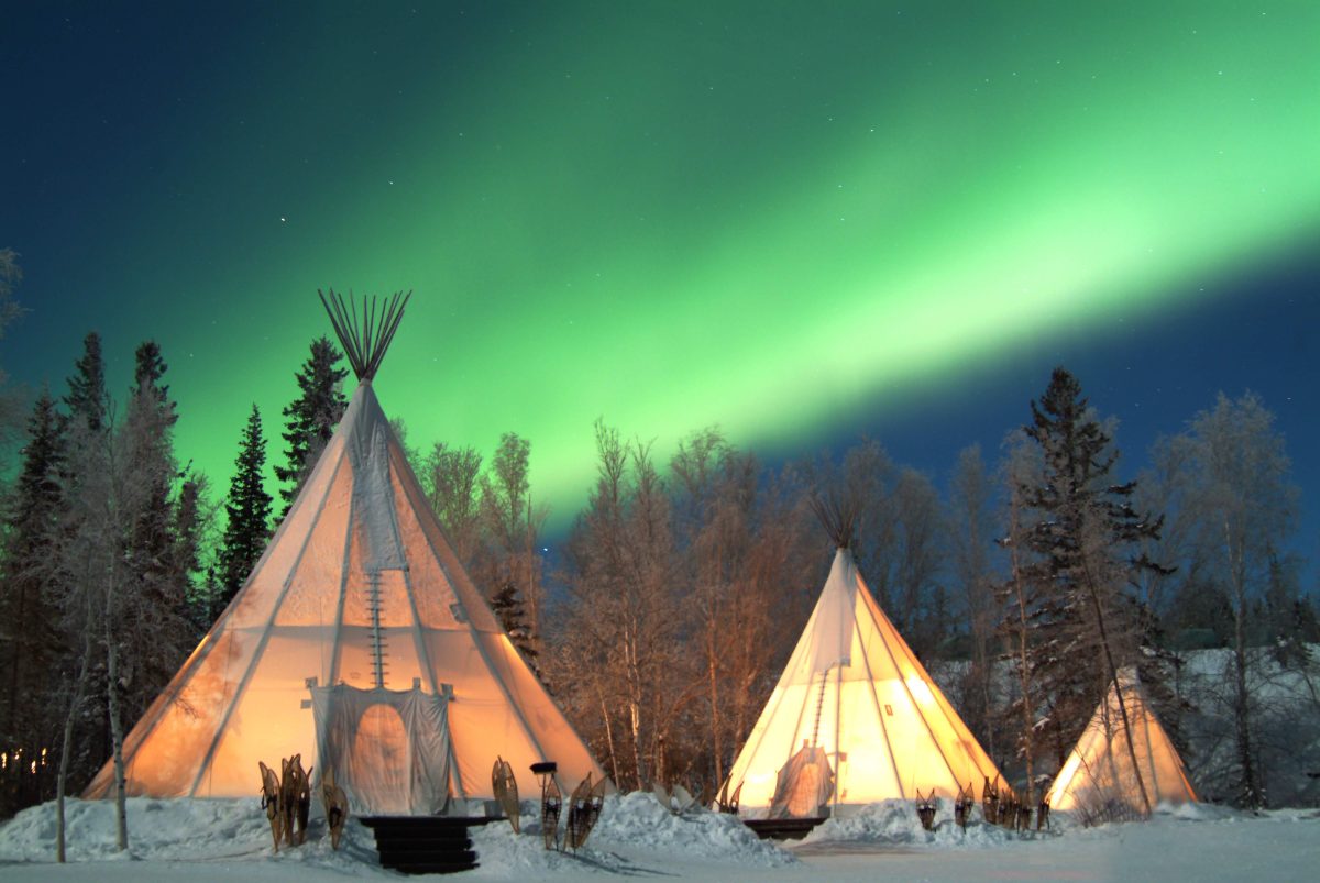 A trio of teepee-style tents with the Northern Lights at in the background at Aurora Village in Yellowknife, one of the many things to experience in Canada during the winter - Luxury Escapes