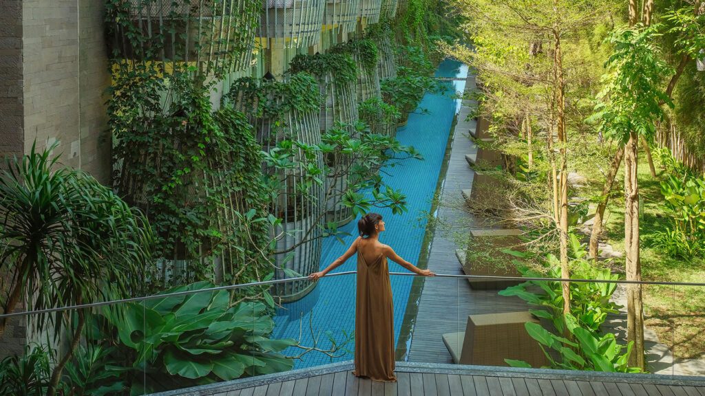 A woman in a brown dress looks over a glass balcony into the gardens of Maya Sanur Resort & Spa which is one of the best family friendly resorts in the world - Luxury Escapes