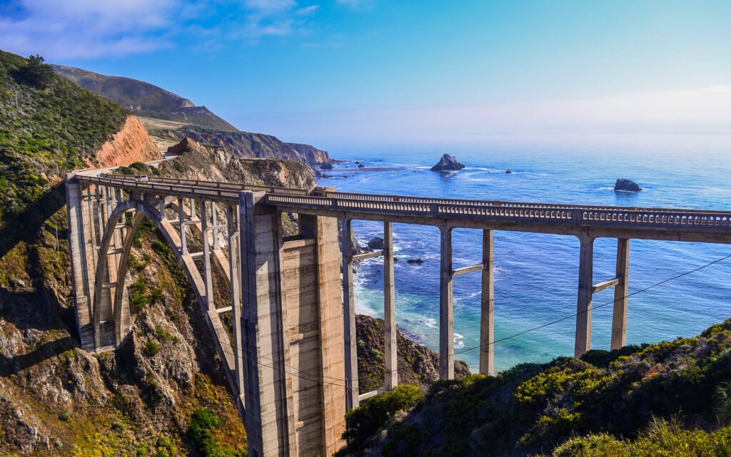 View of the Pacific Coast Highway with the bridge stretching from one side of the cliff to another, and the blue ocean on the horizon for one of the 5 iconic USA road trips - Luxury Escapes 