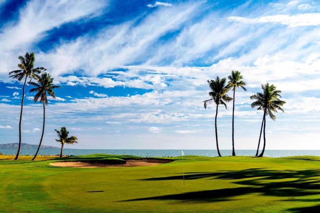 A row of palm trees swaying in the background of this golf green course witht he sea in the background at Sheraton Fiji Golf & Beach Resort, one of the best resorts to stay in Fiji on a weekend retreat from Brisbane - Luxury Escapes