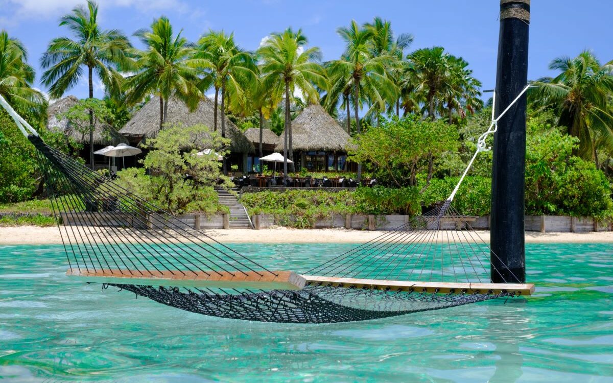 A hammock hanging over the sparkling sea with Likuliku Lagoon Resort in the background, one of Fiji's best all-inclusive resorts - Luxury Escapes