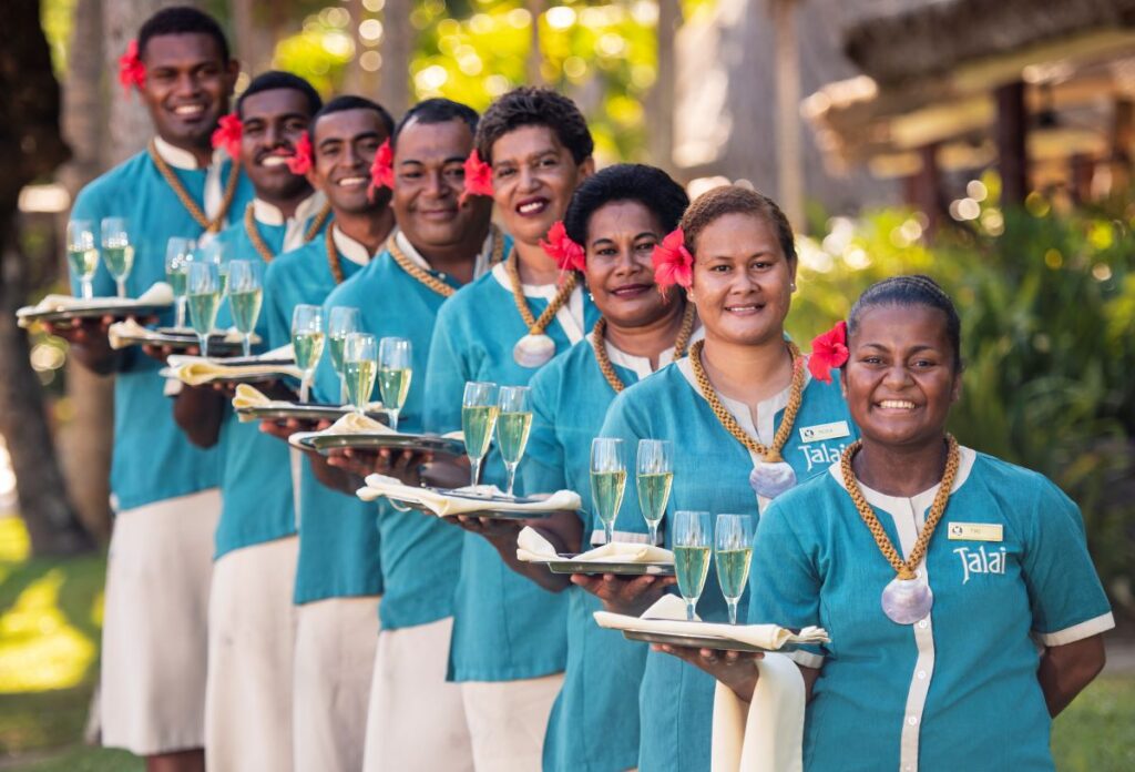 A line of resort staff wearing blue uniforms smile as they hold trays of champagne at Outrigger Fiji Beach Resort, one of the best places to stay in Fiji on a weekend away from Brisbane - Luxury Escapes