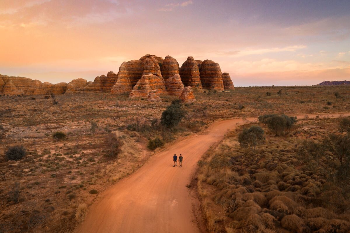 A pair of people walking down a dirt road with a rock formation in the background in the desert in Western Australia - Luxury Escapes
