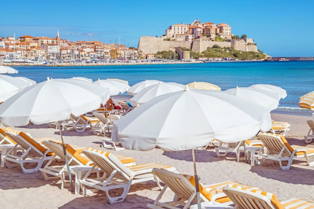 Rows of white umbrellas and sun beds overlooking a beach in Corsica, France, a destination in Europe which is best explored during June - Luxury Escapes