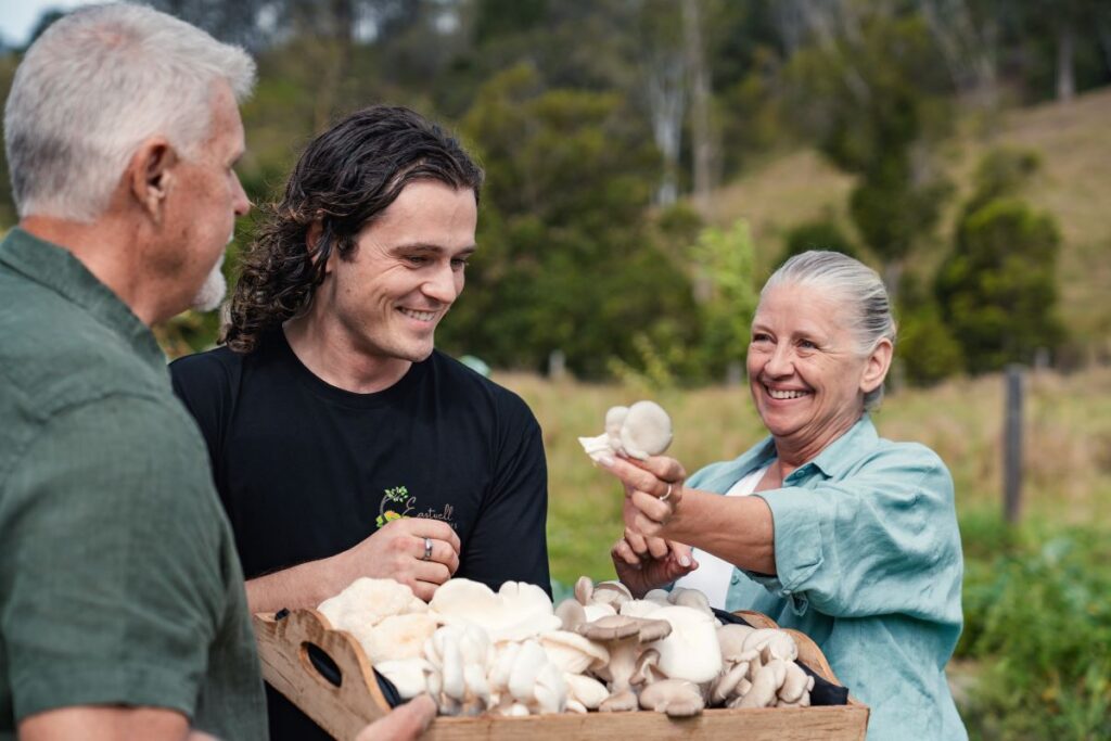 A mushroom farmer extends a mushroom towards two male customers as part of The Curated Plate festival, one of Australia's best food and drink festivals - Luxury Escapes