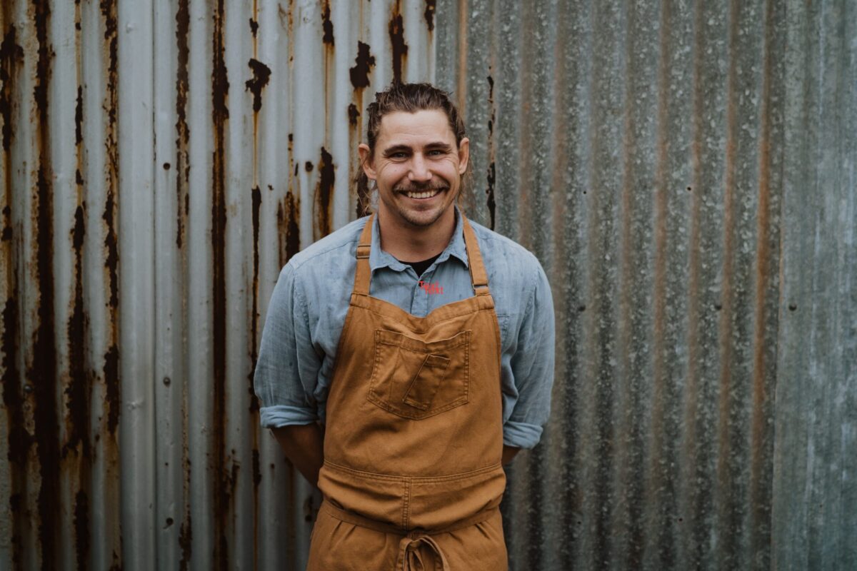 Chef Paul Iskov with a orange apron smiling in front of a corrugated iron fence discussing Pop-Up Restaurants around Western Australia - Luxury Escapes