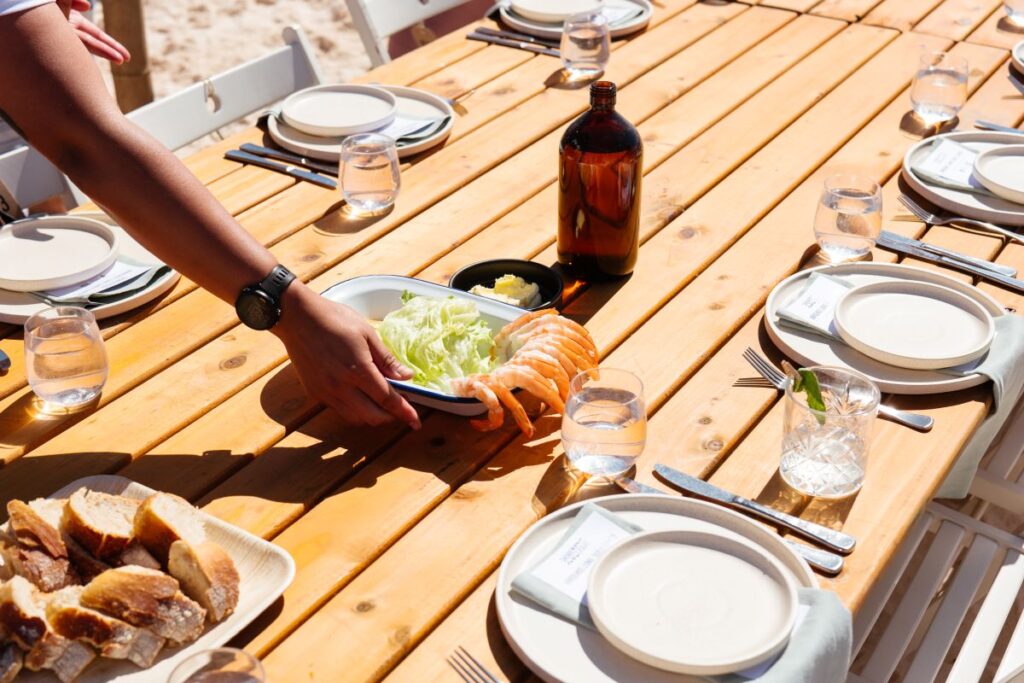 A persons arm places a plate of prawns and lettuce on a set table as part of A Table Somewhere festival, one of Australia's best food and drink festivals - Luxury Escapes