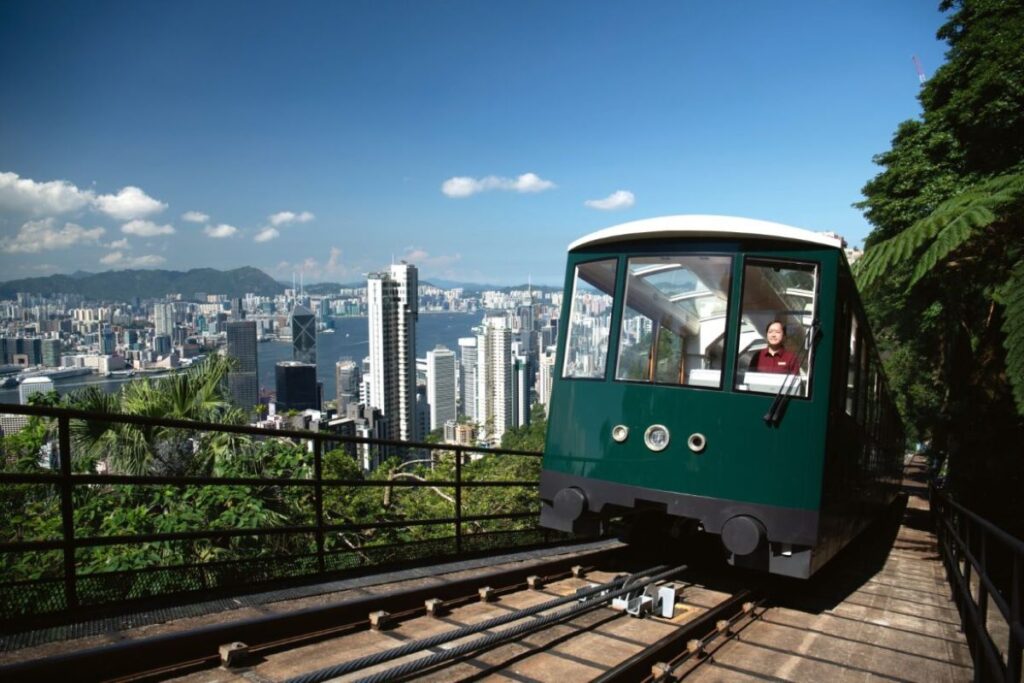 A tram driver drives a Peak Tram up an incline with the Hog Kong skyline in the background which makes the ideal start or ending destination to any cruise in 2024 - Luxury Escapes