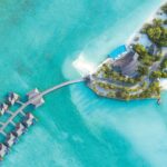 An aerial shot of Hideaway Beach Resort & Spa's floating villas and the blue coastline, one of the best resorts to travel solo in the Maldives - Luxury Escapes