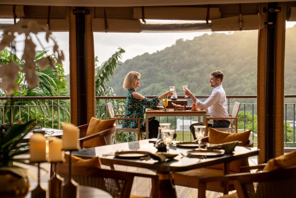 A couple share a cheers overlooking the mountains and ocean of Phuket while dining at one of the city's best restaurants, Tambu - Luxury Escapes