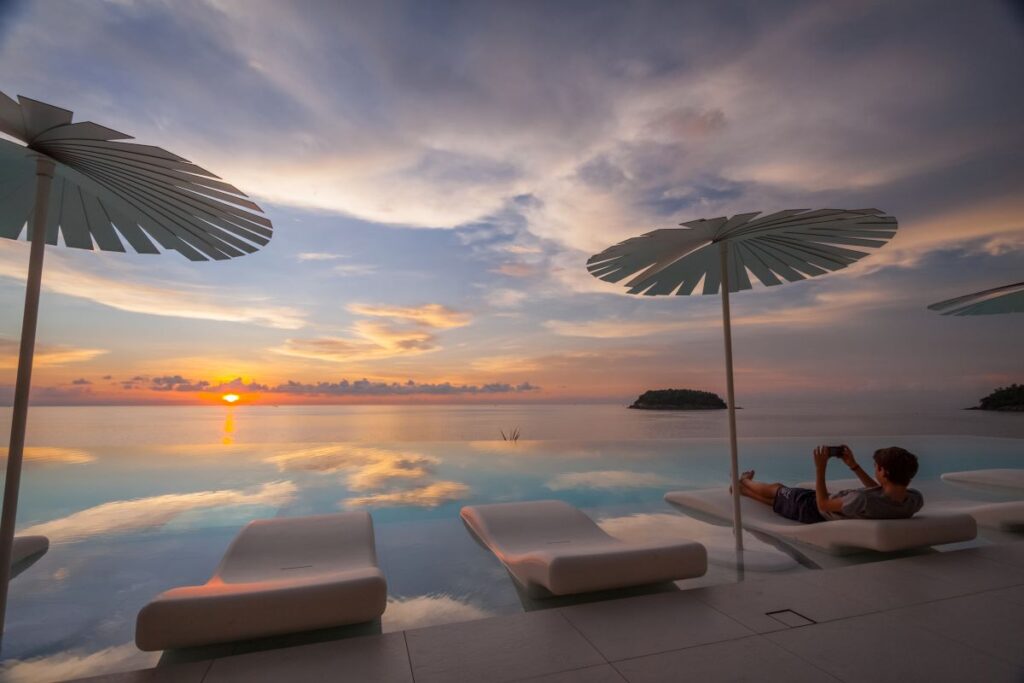 A man reclining poolside taking a photo of the sunset at Kata Rocks, one of Phuket's best resorts - Luxury Escapes