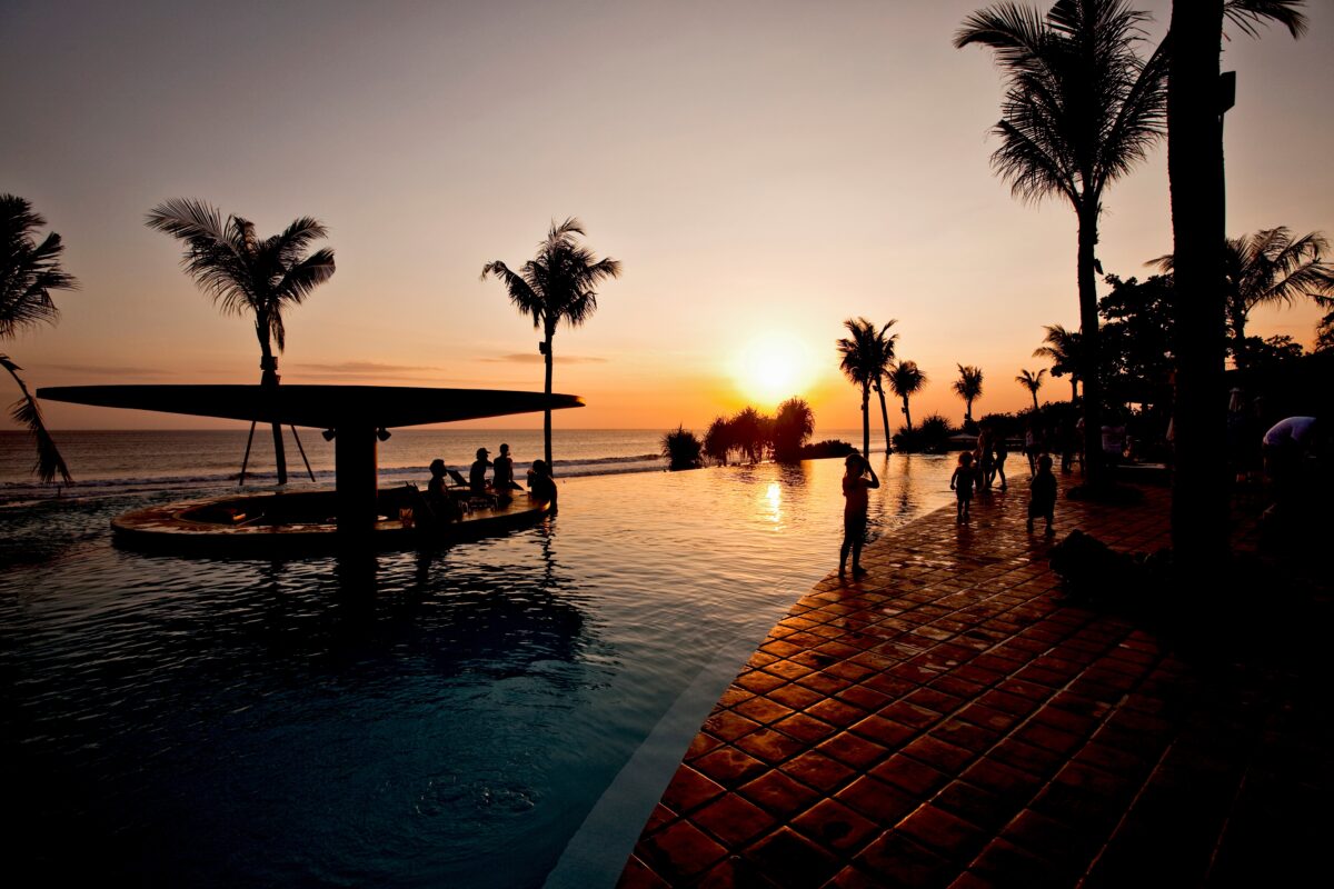 A sunset across the ocean behind a beachfront infinity pool with sunken bar and guests lounging on the terrace beside it.