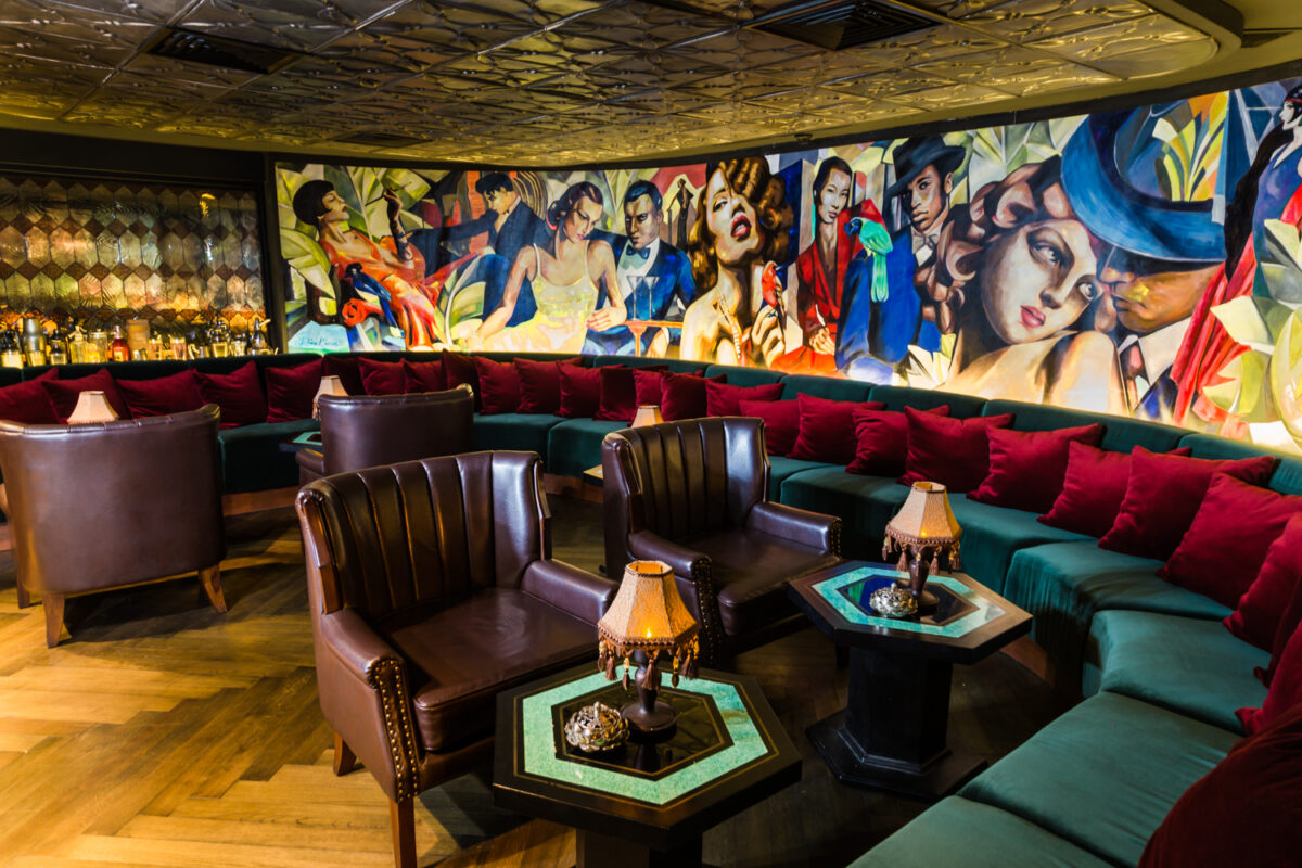 A brightly lit cocktail section of a bar with chairs, banquettes, tables and Art Deco murals.