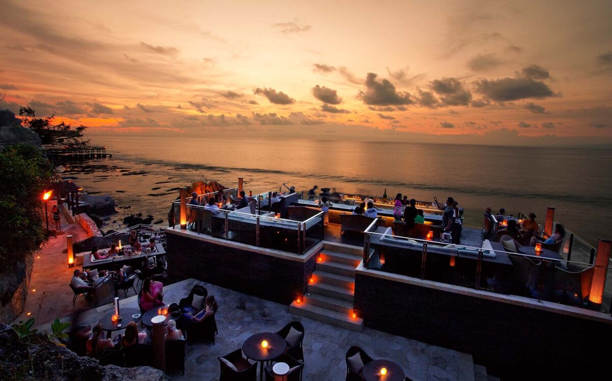 A busy cocktail bar set atop beach cliffs, overlooking a dusky ocean horizon.