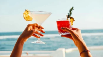 Two hands raising cocktail glasses in front of a backdrop of ocean surf at Tropical Temptation Beach Club.