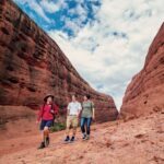 A tour guide walks with two travellers through a red sandstone canyon beneath a blue sky and blanket of white clouds - Luxury Escapes