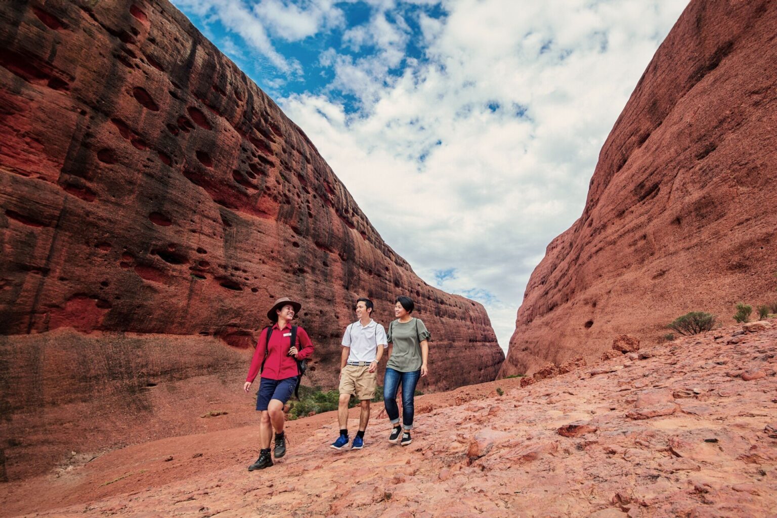 A tour guide walks with two travellers through a red sandstone canyon beneath a blue sky and blanket of white clouds - Luxury Escapes