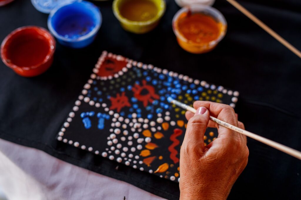 A hand holds a paintbrush over a black canvas covered with a handmade dot painting while red, blue, yellow and orange acrylic paints sit nearby - Luxury Escapes