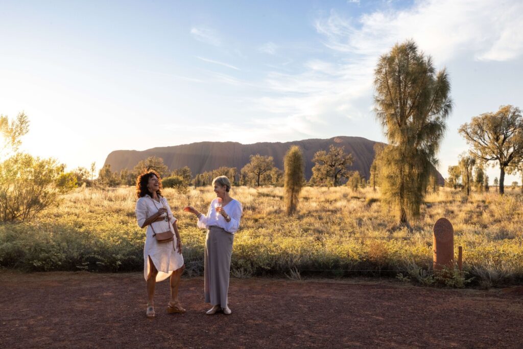 Two women holding glasses of sparkling wine and laughing during sunset in the foreground, while the sandstone monolith, Uluru, falls into shadow in the background - Luxury Escapes