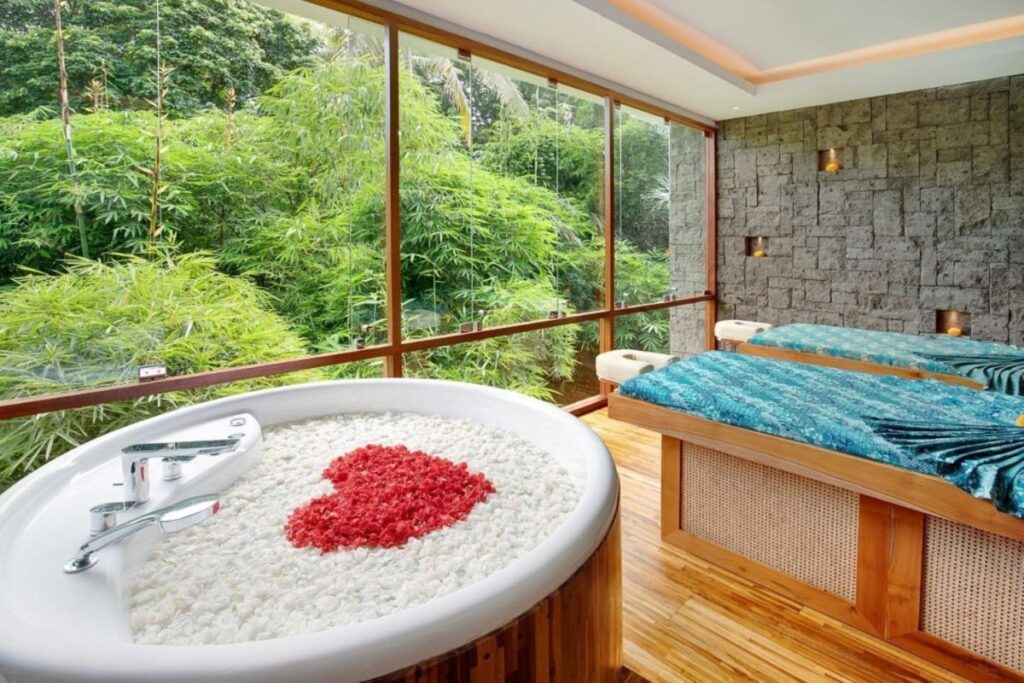 Two massage tables and a tub full of flower decor in a red heart shape sit before floor-to-ceiling windows looking out onto the jungle - Luxury Escapes