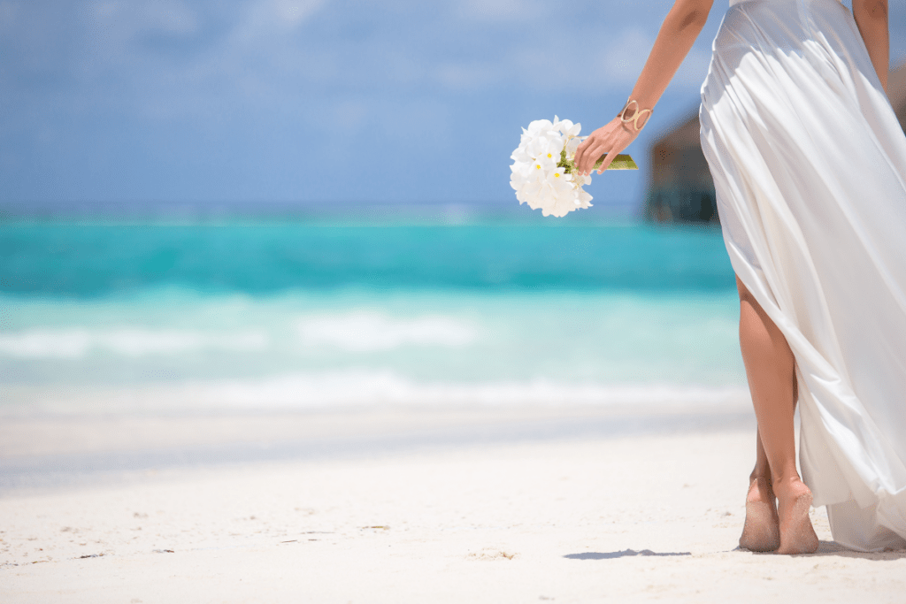 A woman holds a bouqet of white flowers as she heads to a destination wedding on a Maldives beach - Luxury Escapes