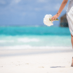 A woman holds a bouqet of white flowers as she heads to a destination wedding on a Maldives beach - Luxury Escapes