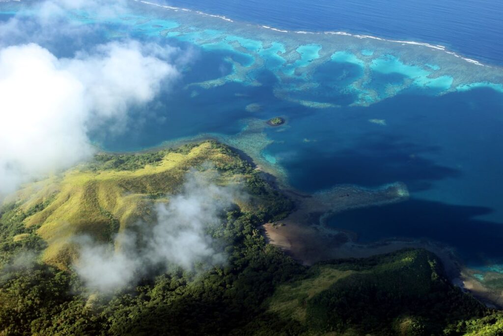 A lush green island with volcanic terrain surrounded by clear blue ocean waters and coral reefs, viewed from above on a partly cloudy day.
