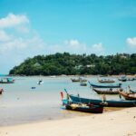 Traditional colorful wooden longtail boats anchored in calm, turquoise waters near a sandy beach on Koh Samui, Thailand, with lush green hills under a bright blue sky.