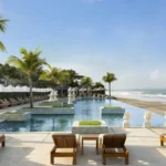 Beachfront Seminyak resort infinity pool with sun loungers, palm trees and ocean views. - Luxury Escapes